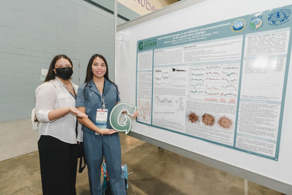 Two women smiling next to a large scientific poster. Both are wearing lanyards with name tags and one holds a large cutout letter "G" for University of Guam