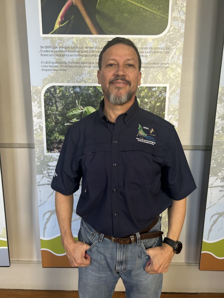 Milton Muñoz wearing a buttondown shirt and jeans stands in front of an educational sign in Jobos Bay