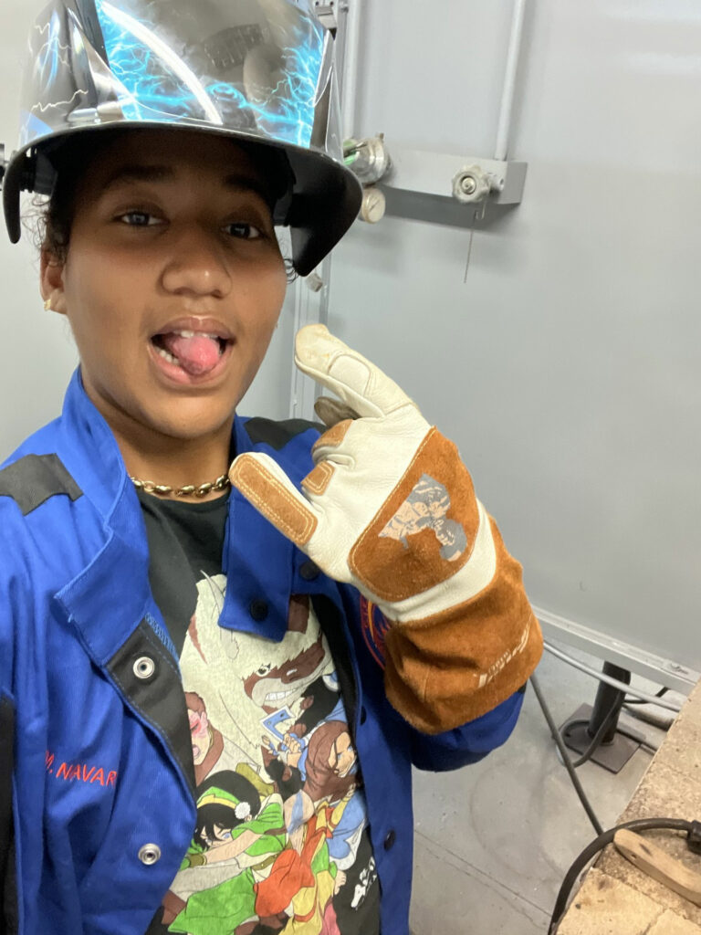 A young woman wearing a welding mask and gloves sticks her tongue out and takes a selfie